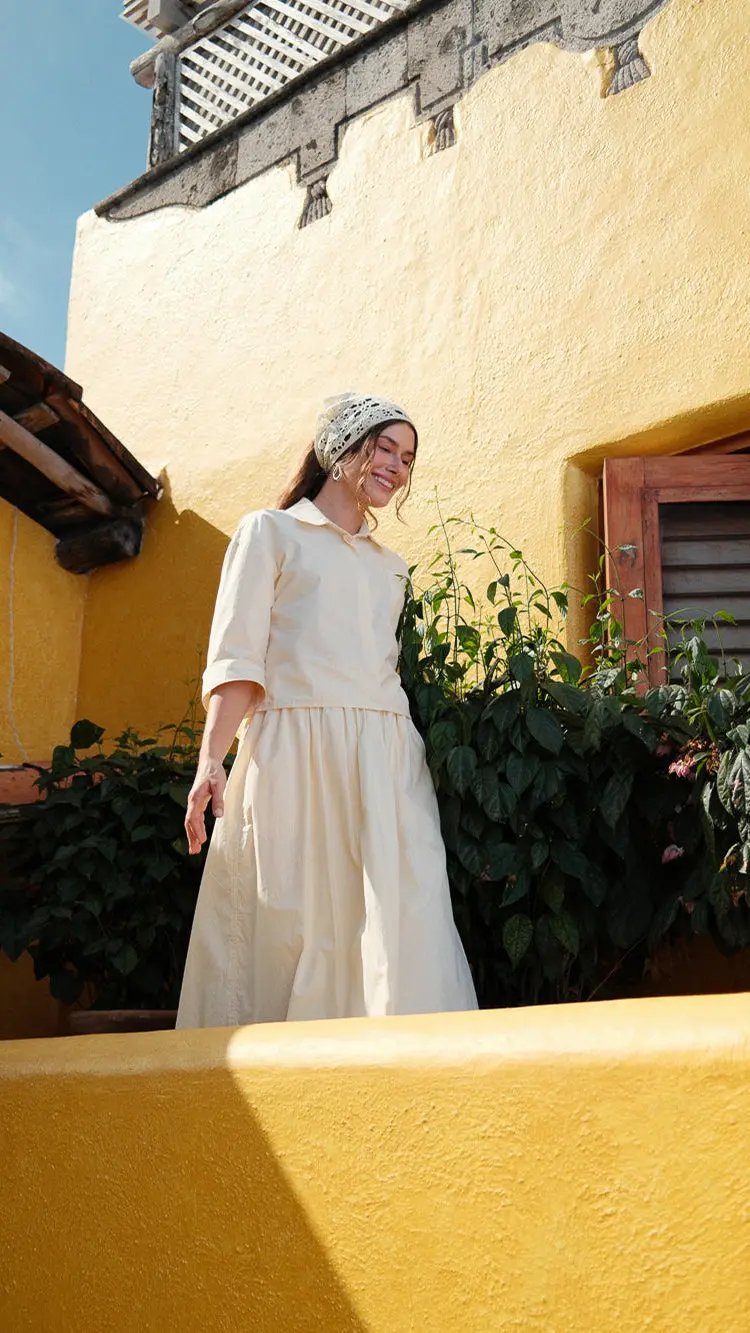 Woman in a white dress standing on a yellow balcony with a yellow wall and plants in the background.